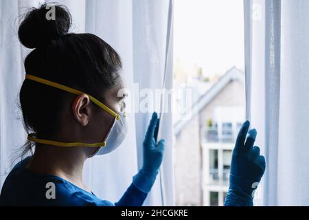 Annoiato donna in quarantena corona con maschera di protezione fftp2 che guarda da una finestra al villaggio Foto Stock