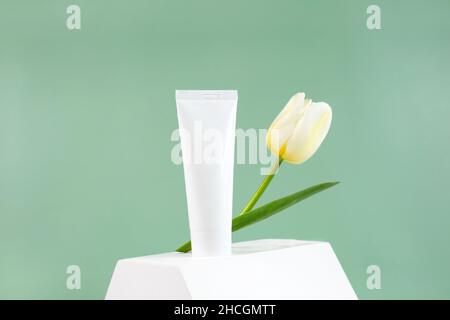 Tubo cosmetico bianco con fiore di tulipano sul podio bianco su sfondo verde. Concetto di minimalismo nell'imballaggio cosmetico. Spazio di copia. Natural Organic Spa Cosmetic Beauty Concept Mockup. Foto Stock