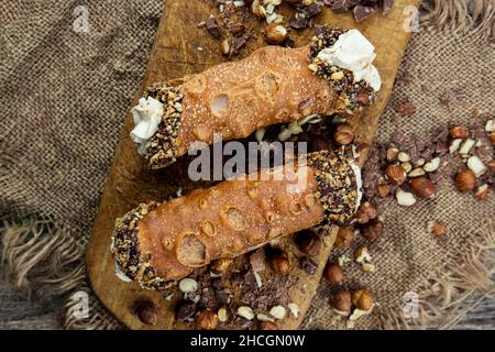 Cannoli, deliziosi tubi di pasta fritta italiana con ricotta dolce, patatine al cioccolato e nocciole serviti su un asse di legno Foto Stock