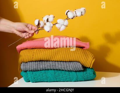 pila di maglioni piegati in maglia e una mano della donna che tiene un ramo di cotone su uno sfondo giallo Foto Stock