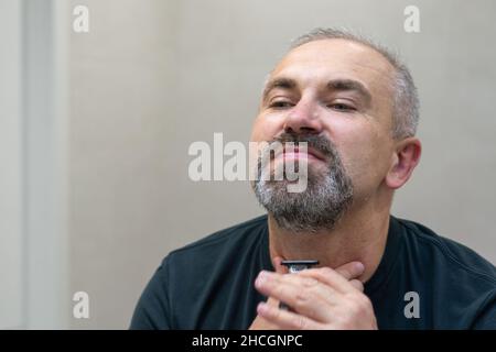 Uomo maturo dai capelli grigi che restyling la sua barba in casa utilizzando il rasoio Foto Stock