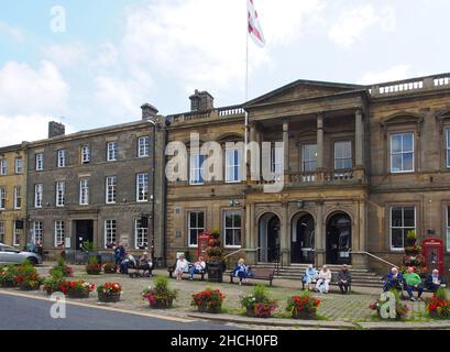 Gli anziani godono del sole seduto su panchine fuori dal municipio di Skipton, North Yorkshire, Inghilterra, con fiori in vasche in primo piano. Foto Stock
