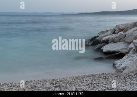 Mare Adriatico, spiaggia di ciottoli e rocce, vane silhouette di isole in lontananza al crepuscolo Foto Stock