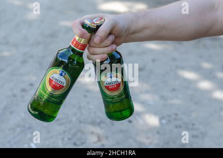 Una mano tiene due bottiglie di birra Amstel contro la sabbia. Maschio adulto con bottiglie verdi di bevanda alcolica. Nikolaev, Ucraina - 07 13 2021 Foto Stock