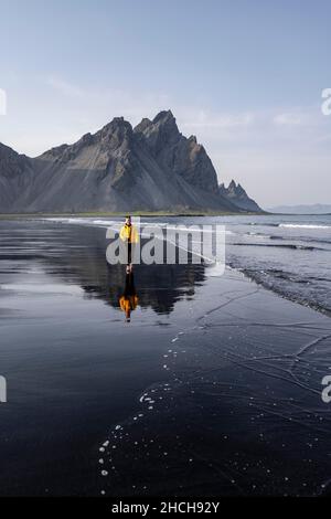 Giovane donna con giacca a pioggia che cammina sulla spiaggia, spiaggia di lava nera, spiaggia sabbiosa, promontorio di Stokksnes, Austurland, Islanda orientale, Islanda Foto Stock