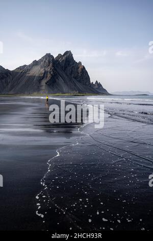 Giovane donna con giacca a pioggia che cammina sulla spiaggia, spiaggia di lava nera, spiaggia sabbiosa, promontorio di Stokksnes, Austurland, Islanda orientale, Islanda Foto Stock