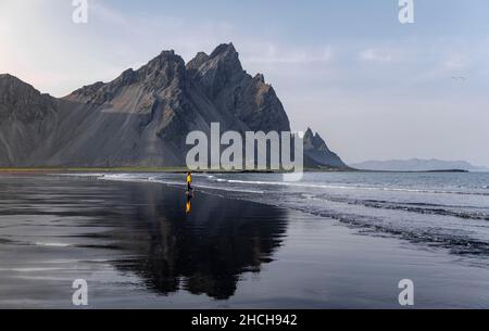 Giovane donna con giacca a pioggia che cammina sulla spiaggia, spiaggia di lava nera, spiaggia sabbiosa, promontorio di Stokksnes, Austurland, Islanda orientale, Islanda Foto Stock