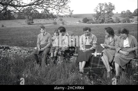 1950s, storica, fuori in un campo di campagna, una famiglia contadina seduta insieme su un muro di pietra con una tazza di tè e uno spuntino, Inghilterra, Regno Unito. Foto Stock