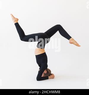 Una giovane donna in abbigliamento sportivo nero, esegue una variante dell'esercizio shirshasana, si allena in studio su uno sfondo chiaro Foto Stock