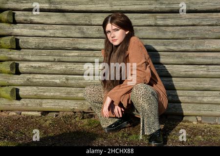 Giovane ragazza si accoccola sul pavimento di fronte a vecchi assi di legno al sole e guarda avanti Foto Stock