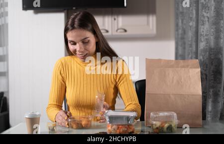 Una bella ragazza caucasica è seduta al tavolo a casa da sola la sera e sta andando a cena da piatti usa e getta. Gambe di pollo fritte Foto Stock