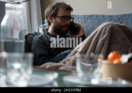 Un giovane uomo, con bicchieri di pasta, bearded, parla al telefono cellulare con qualcuno, mentre si riposa sul divano, coperto da una coperta, nell'apa condiviso Foto Stock