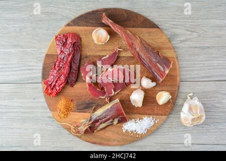 Carne di maiale affumicata a fette su tagliere di legno scuro rotondo, organizzato con pepe rosso secco, aglio e pepe di cayenna su sfondo grigio di legno, top Foto Stock