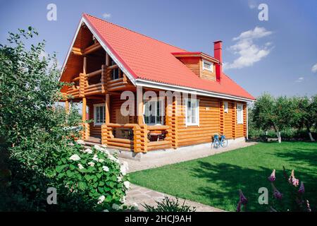 Grande casa di campagna in legno di recente costruzione con tetto di tegole rosse, prato sulla destra e frutteto di alberi di ciliegio dietro casa e giardino fiorito anteriore in Foto Stock