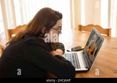 Bella donna latina seduta nella sala da pranzo della sua casa durante il giorno vicino al suo laptop che parla con il suo amico su una videochiamata Foto Stock