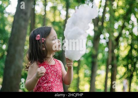 Piccola ragazza carina 3-4 mangiare caramelle di cotone in parco soleggiato tra alberi alti su erba verde Foto Stock