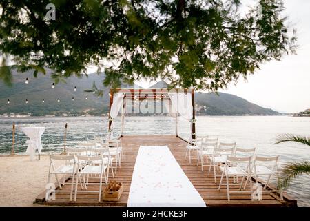 File di sedie bianche si trovano sul molo di fronte all'arco del matrimonio sullo sfondo delle montagne Foto Stock