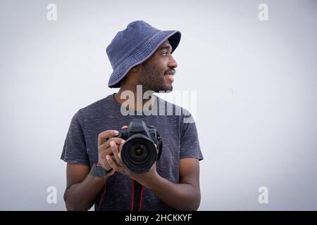 Il fotografo professionista tiene una fotocamera in mano. L'uomo elegante e attraente indossa un cappello da secchio e sorride mentre tiene una DSLR, è isolato su Whi Foto Stock