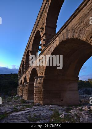 Vista laterale ad angolo basso dell'antico acquedotto romano Pont du Gard con maestosi archi in pietra sopra il fiume Gardon dopo il tramonto vicino a Vers-Pont-du-Gard, Francia. Foto Stock