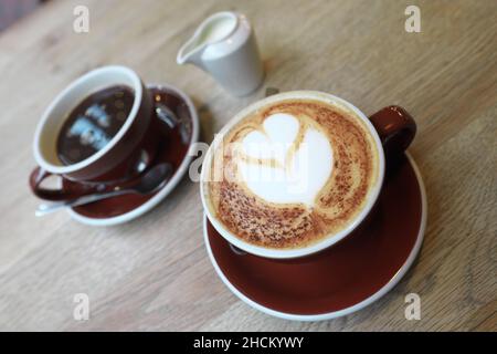 Un caffè cappuccino con arte del caffè decorata con design a cuore e un caffè nero con caraffa di latte dietro Foto Stock