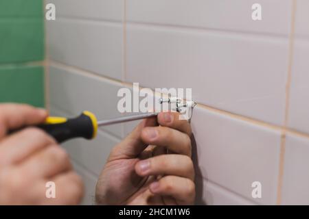 Uomo mani con cacciavite avvitando chiodi in tasselli martellato in parete piastrellata in bagno. Lavori di ristrutturazione nella stanza da bagno. Sporcizia dal processo di ristrutturazione Foto Stock