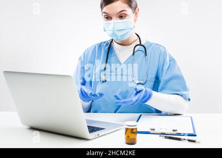 Giovane donna caucasica NHS GP medico seduto alla scrivania in ufficio, indossare uniforme blu, maschera protettiva e guanti, visita virtuale tele paziente Foto Stock