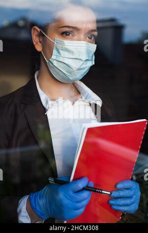 Giovane donna d'affari con espressione facciale preoccupata, indossare maschera protettiva, tenere cartella rossa file, guardando attraverso la finestra in un futuro incerto Foto Stock