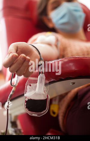Giovane donna che dona sangue in ospedale o laboratorio, seduta in sedia con sacco di sangue a fuoco, convalescente COVID-19 paziente trasfusione plasma Foto Stock