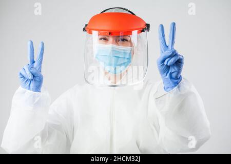 Giovane tecnico medico che indossa i dispositivi di protezione individuale PPE, tuta bianca, visiera protettiva, maschera e guanti, festeggiamenti mostrando il segno del dito V. Foto Stock