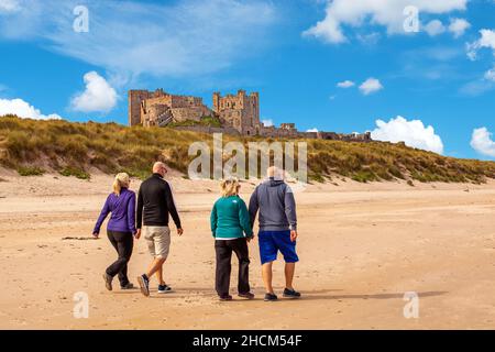 Due coppie camminare tenendo le mani lungo la spiaggia sabbiosa di Northumberland Bamburgh England Regno Unito con il castello di Bamburgh in background Foto Stock