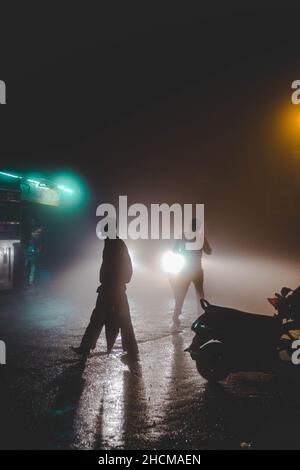 Silhouette di persone che camminano lungo la strada con luci per la strada su di esso coperto di notte nella nebbia Foto Stock