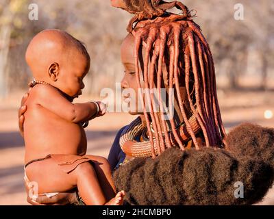 Palmwag, Namibia - 21 agosto 2016. Una giovane donna africana tiene il suo bambino mentre indossa il tradizionale hairstyle e pasta di pelle ocra della tribù Himba. Foto Stock