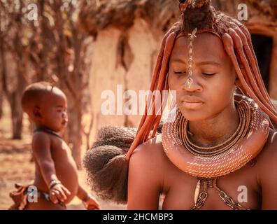 Una donna africana che indossa gioielli tribali e acconciature tradizionali Himba con il suo bambino in background nella Namibia rurale. Foto Stock