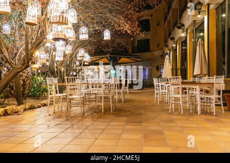 Terrazza dell'hotel in un cortile vuoto di notte con lanterne illuminate negli alberi, tavoli sui lati e un bar sullo sfondo Foto Stock