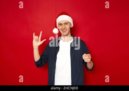 Sorridente giovane uomo felice in abiti casual con cappello di Capodanno mostra segno di sordi e muto con la sua mano ti amo isolato su sfondo rosso per il concetto di testo - salute, comunicazione, simpatia. Foto di alta qualità Foto Stock