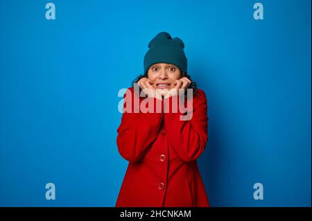 Giovane donna bruna dai capelli ricci in cappotto rosso brillante e cappello verde con uno sguardo spaventato guardando la macchina fotografica, grattando i denti, tenendo la mano su h Foto Stock