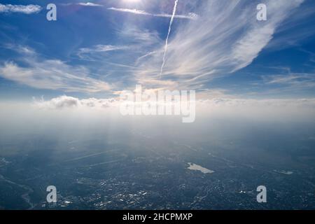 Vista aerea dalla finestra dell'aeroplano ad alta quota di città lontana coperta da strato di smog sottile e nubi distanti. Foto Stock