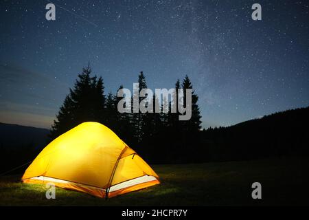 Luminosa tenda turistica illuminata che risplenderà sul campeggio in montagne scure sotto il cielo notturno con stelle scintillanti. Concetto di stile di vita attivo. Foto Stock