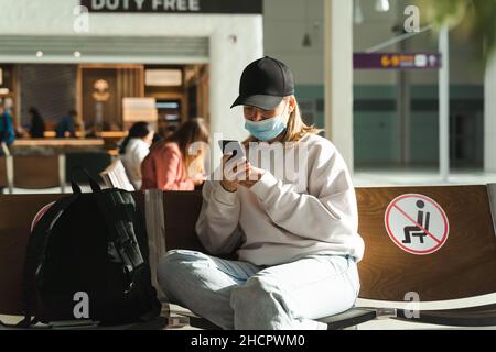 Giovane ragazza che indossa una maschera medica e usa lo smartphone seduto nella lounge del terminal dell'aeroporto in attesa di partenza. Viaggio durante la quarantena Foto Stock