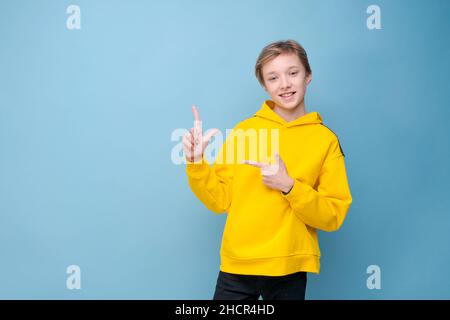 Felice allegro sorridente giovane caucasico con capelli biondi, indossando una felpa gialla su sfondo blu, guardando verso l'alto e puntando verso l'alto con il dito indice. Spazio di copia e concetto di annuncio. Foto Stock