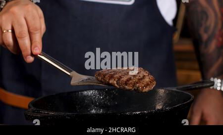 Primo piano dello chef maschile friggere la cotoletta di hamburger nella padella nera di ferro da entrambi i lati con l'aiuto di una pala. Uomo in grembiule preparare la cotoletta di carne. Foto Stock