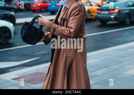 Abito femminile alla moda in autunno. Donna elegante che cammina in cappotto marrone con borsa nera all'aperto. Abbigliamento moderno alla moda. Moda Street style Foto Stock