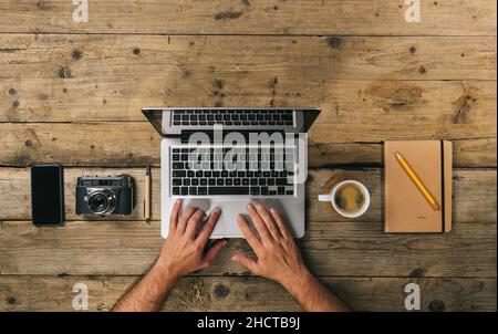 Fotografo che lavora alla scrivania dell'ufficio con computer portatile, notebook argento, telefono cellulare, penna, macchina fotografica e tazza di caffè, piatto. Vista dall'alto con copyspace per il Foto Stock