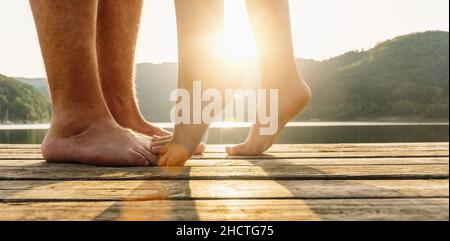 Gambe sul molo. Una giovane coppia affettuosa abbraccia e bacia sul molo su un lago al tramonto. Due amanti, uomo e donna a piedi nudi vicino all'acqua su un molo Foto Stock