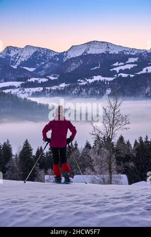 Paesaggio con donna che guarda l'alba sulla catena montuosa di Nagelfluh durante un'escursione con racchette da neve nelle Alpi di Allgaeu vicino a Oberstaufen, Germania, Baviera, Foto Stock