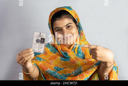 Una giovane donna indiana sorridente e che punta alla carta elettore in mano su sfondo bianco Foto Stock