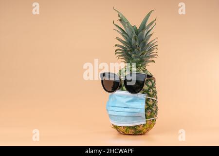 Interpretazione della frutta di ananas in forma umana durante una pandemica indossare occhiali da sole e una maschera di protezione medico. Spazio di copia su sfondo giallo Foto Stock