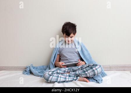 Un bel ragazzo, un adolescente in pigiama grigia, sul pavimento sorride, usa un tablet digitale Foto Stock