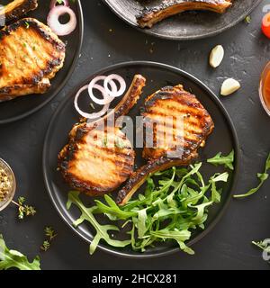 Carne di maiale alla griglia bistecche sulla piastra sulla pietra nera dello sfondo. Vista superiore, laici piatta Foto Stock