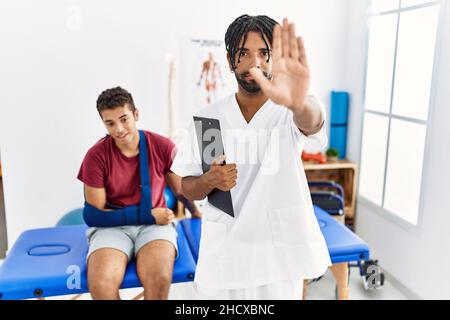 Giovane ispanico che lavora alla clinica di recupero del dolore con un uomo con braccio rotto che fa smettere di cantare con il palmo della mano. Avvertimento espressione con negativo Foto Stock